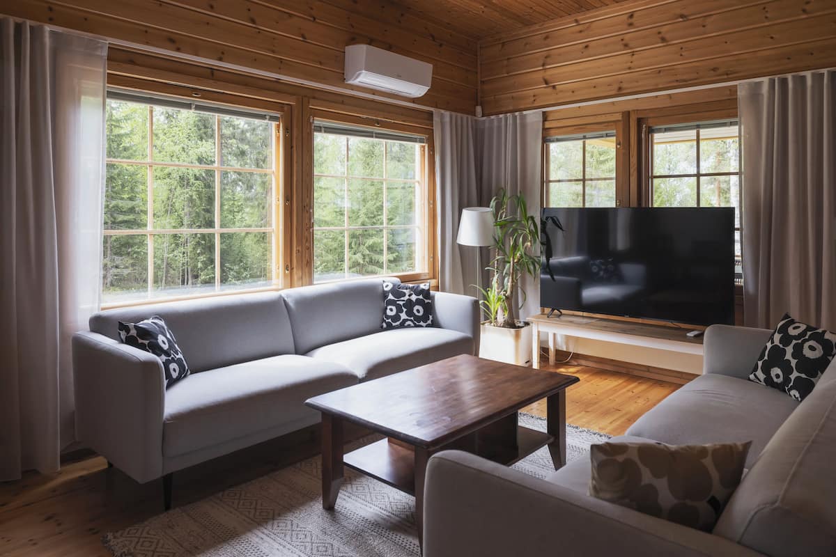 Una sala de estar acogedora con un sofá, una mesa de centro y un televisor. Hay ventanas grandes con cortinas, una lámpara y una planta.