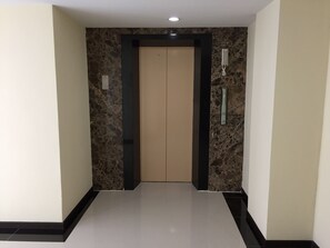 Un pasillo con pared de mármol, puerta de ascensor color beige y zócalo negro.