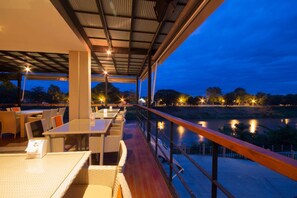 Un espace extérieur aménagé pour les repas, avec des tables et des chaises, offrant une vue sur une étendue d’eau au crépuscule.