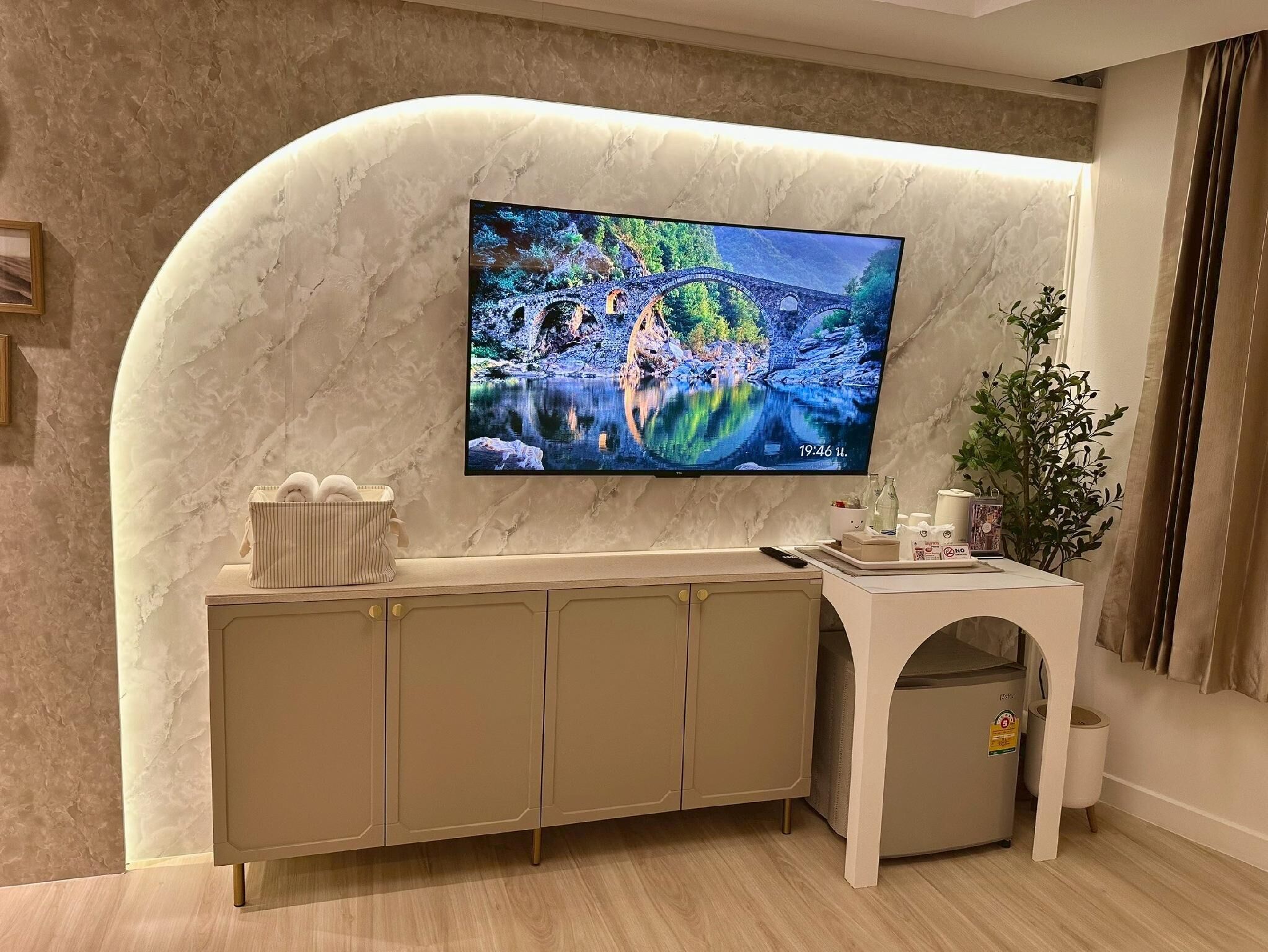 A modern living room with a TV mounted on a curved wall, a wooden cabinet, a small table with a plant, and a trash bin.