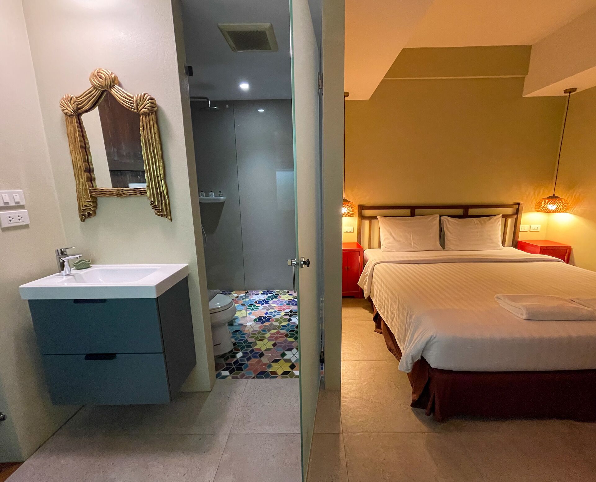 A hotel room with a bathroom, a shower, a bed with white bedding, and a red nightstand.