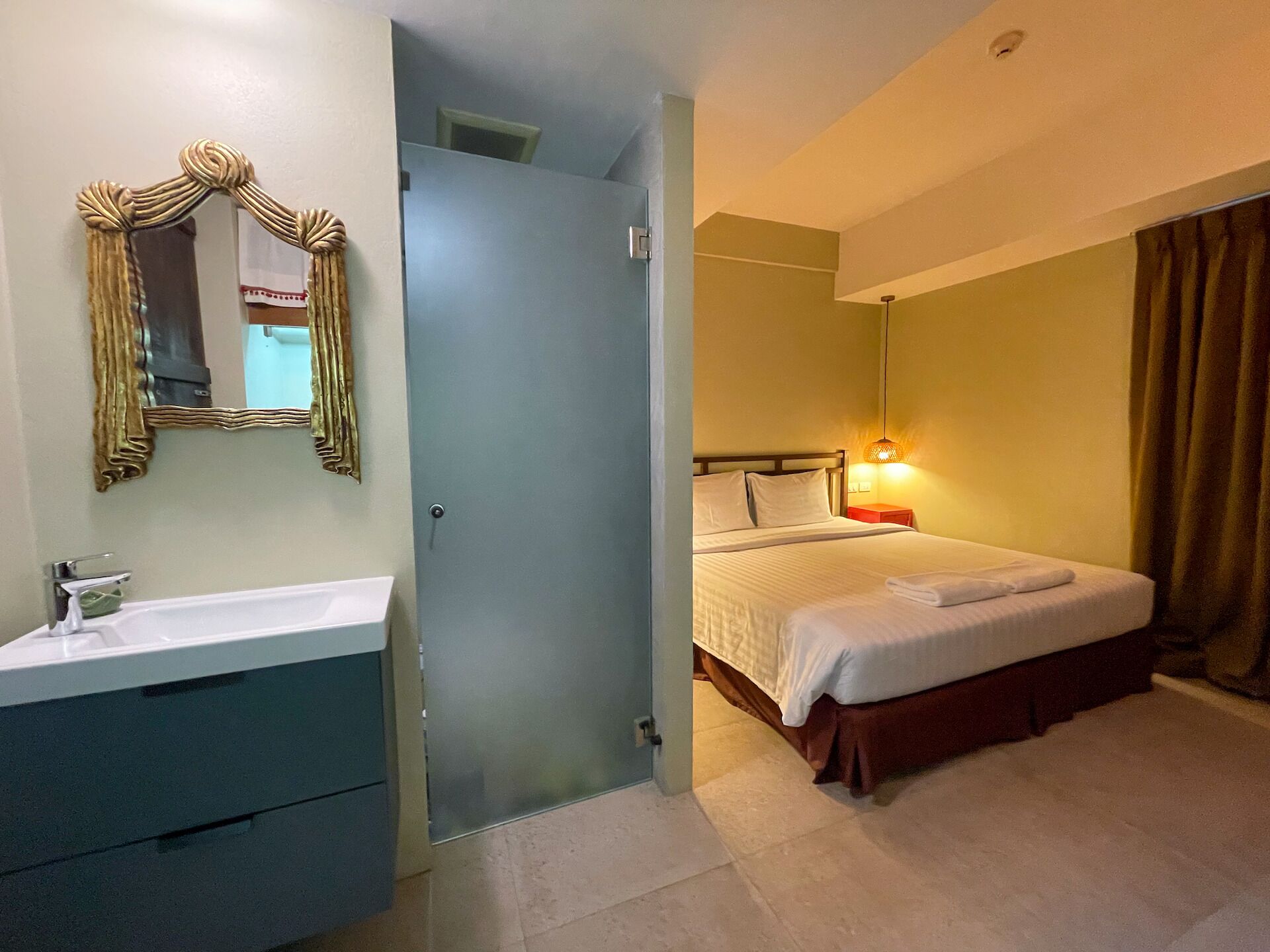 Habitación de hotel con cama, mesita de noche, lámpara y baño con lavamanos y espejo.