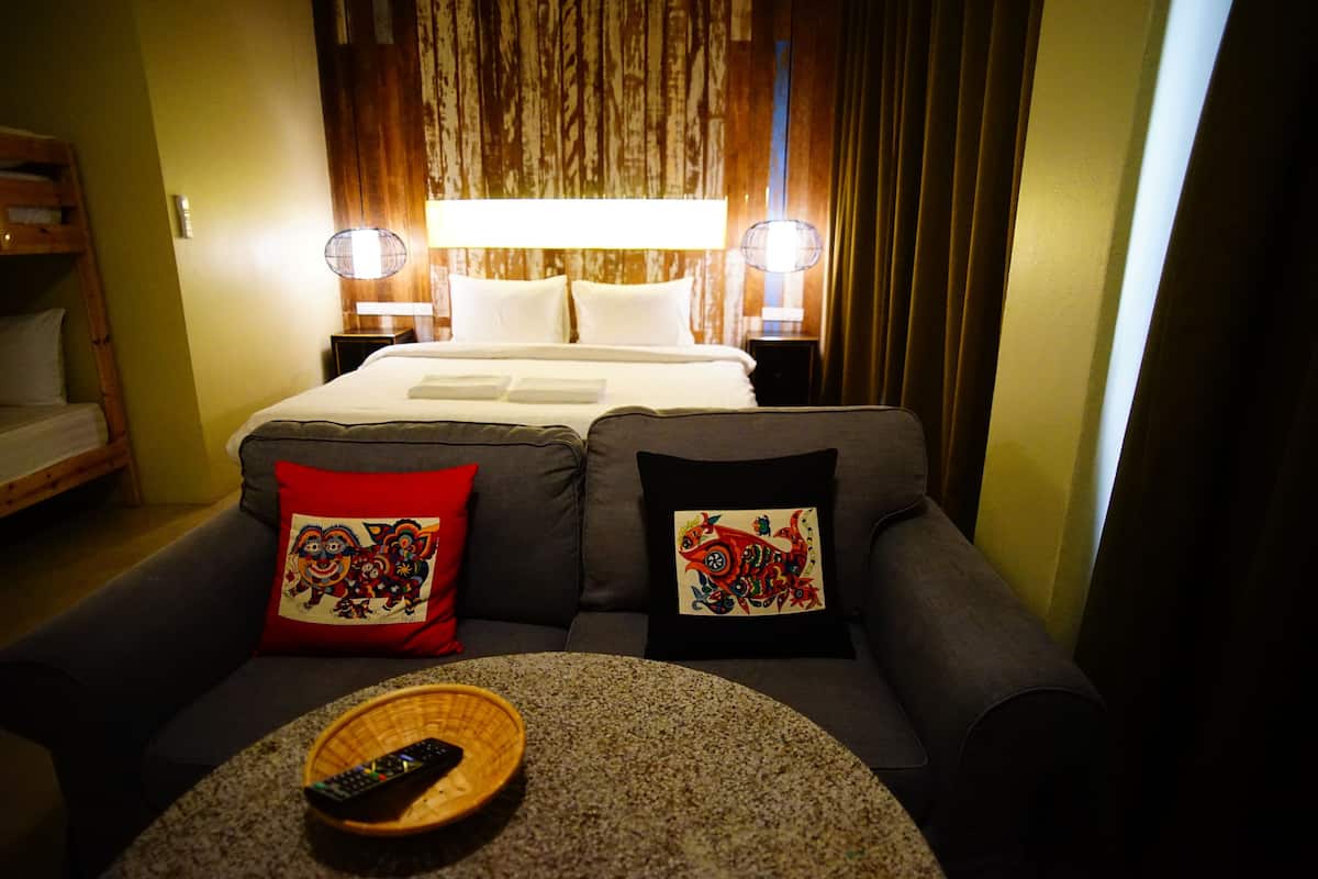 A hotel room with a bed, two bedside lamps, a sofa with decorative pillows, and a remote control on a round table.