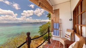 Een balkon met uitzicht op turquoise water en verre bergen, een rieten stoel en een gestreepte handdoek aan de muur.