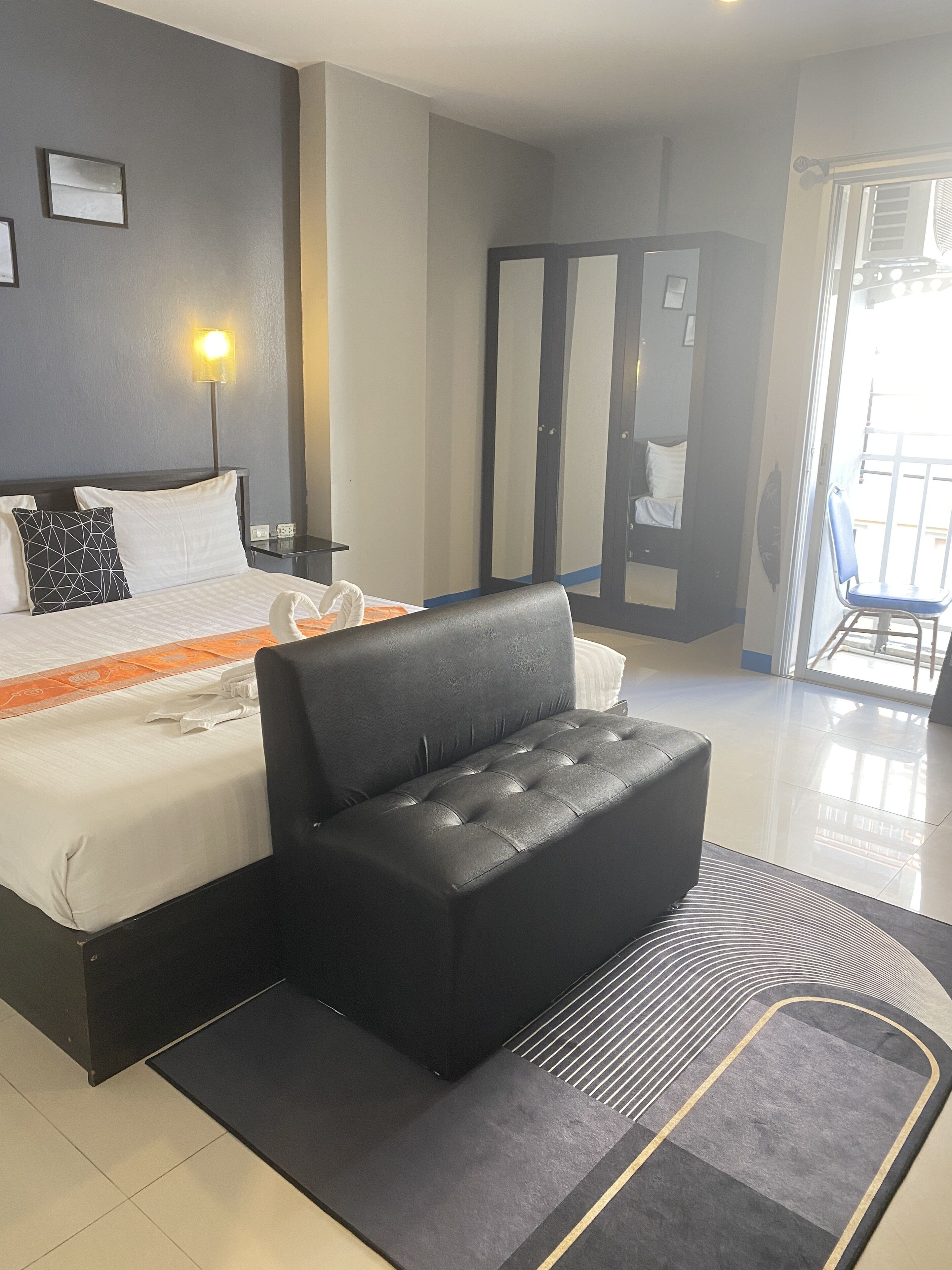 A modern hotel room with a large bed, a black sofa, a glass door leading to a balcony, and a nightstand with a lamp.