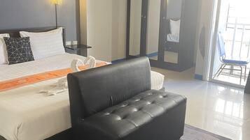 A modern hotel room with a large bed, a black sofa, a glass door leading to a balcony, and a nightstand with a lamp.
