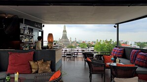 Ein Sitzbereich im Freien mit Korbmöbeln, eine Bar mit Flaschenregalen und ein Blick auf einen Tempel im Hintergrund.