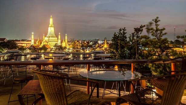 Eine Dachterrasse mit Tischen und Stühlen, die auf eine städtische Szene im Abenddämmerung blickt, mit beleuchteten Gebäuden und einem markanten Tempel.