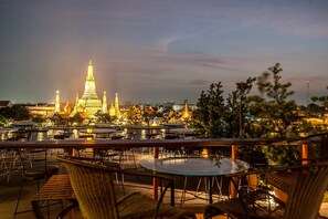 A rooftop terrace with tables and chairs overlooking a cityscape at dusk, featuring illuminated buildings and a prominent temple. - Sala Arun (Bangkok)