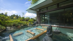 A clear outdoor pool with wooden logs and rocks, surrounded by lush greenery and a modern building with large windows.