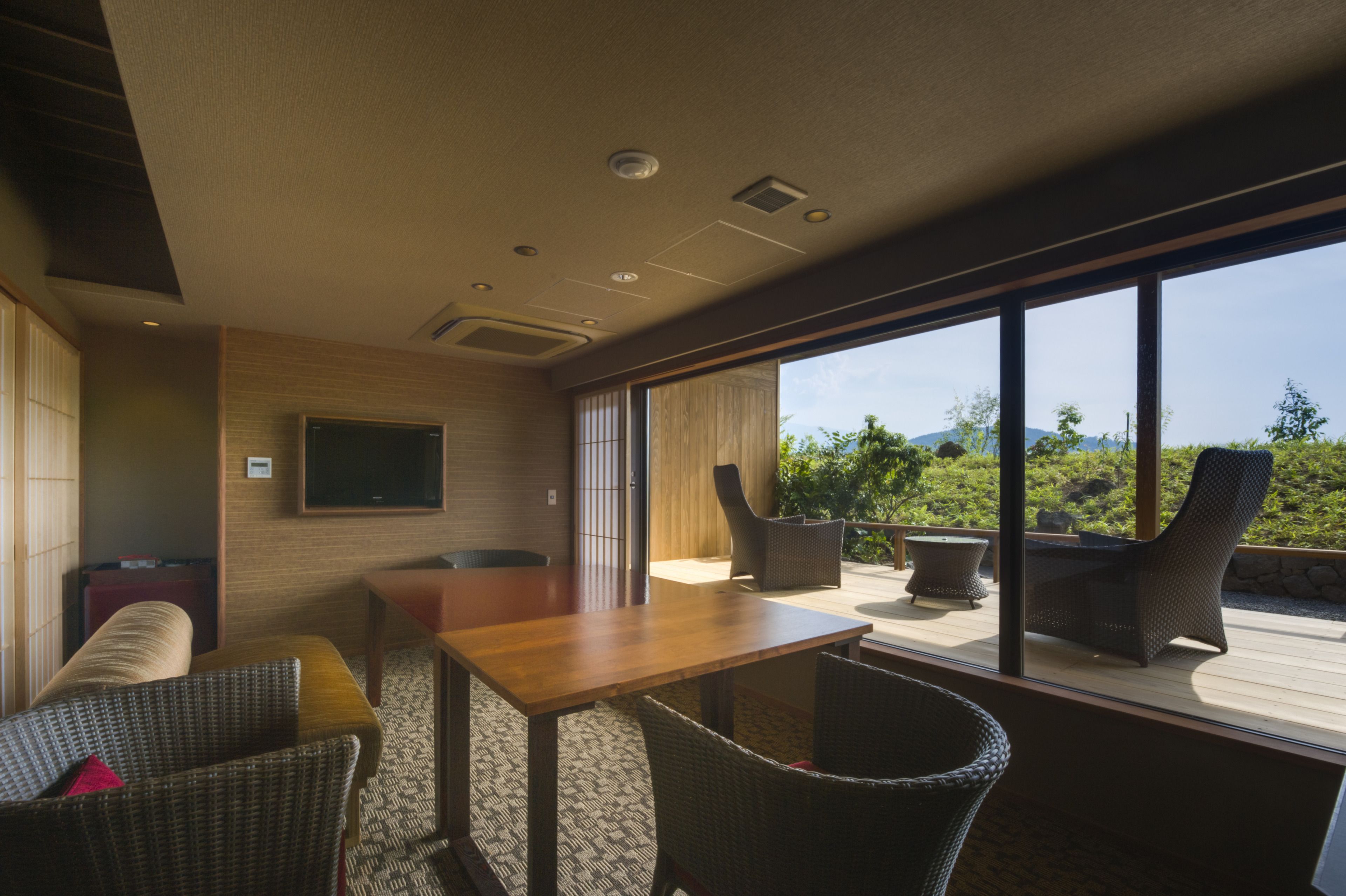 A modern interior with a wooden table and wicker chairs, a flat-screen TV mounted on the wall, and a view of greenery outside.