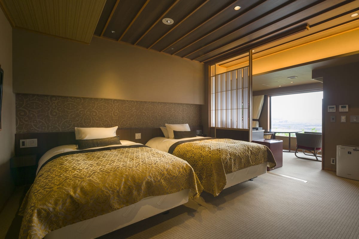 Ein Hotelzimmer mit zwei Betten, einem großen Fenster, einem Schreibtisch und Blick auf die Stadt.
