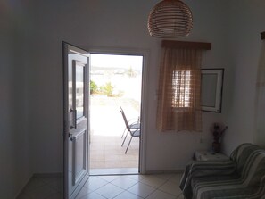 A room with a door leading to an outdoor area, a window with sheer curtains, a chair outside, and a sofa inside. - Kavousanos Apartments (Agios Nikolaos)