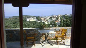 A patio with two wooden chairs and a table, overlooking a coastal town with buildings and greenery.