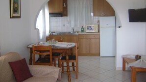 A compact kitchen with wooden cabinets, a microwave, and a refrigerator. There is a dining table with chairs and a sofa in the living area.