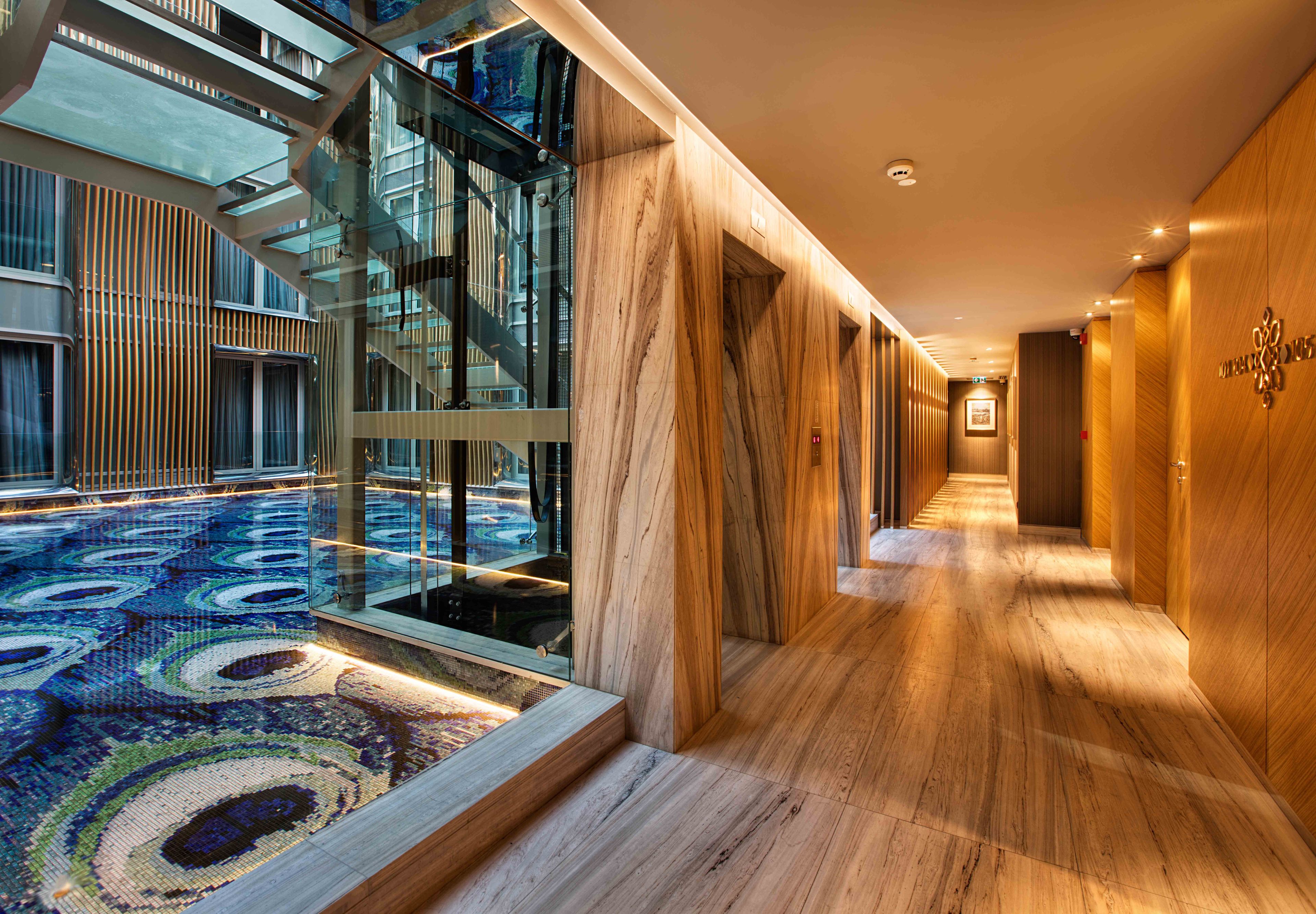A modern hotel corridor with a glass elevator, wooden paneling, and a patterned carpet.