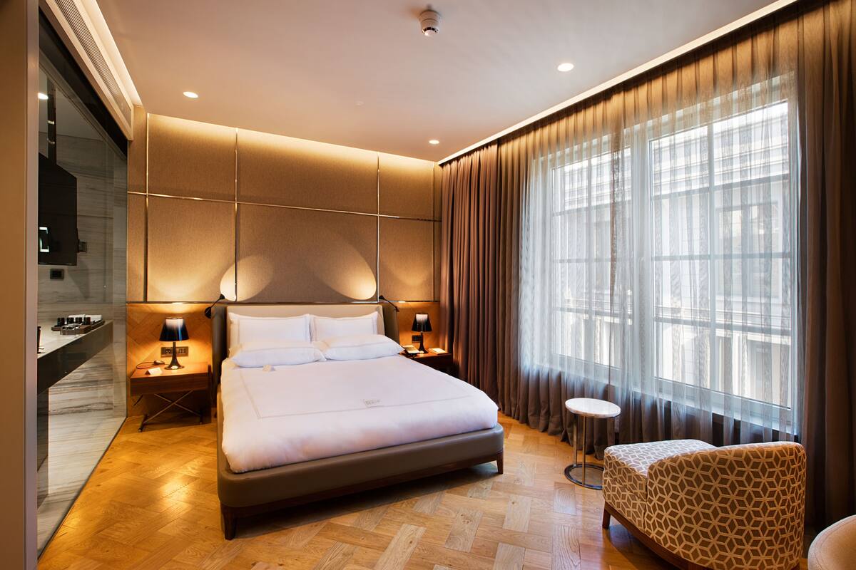 Une chambre d’hôtel moderne avec un grand lit, des lampes de chevet, une petite table ronde et un fauteuil à motifs.