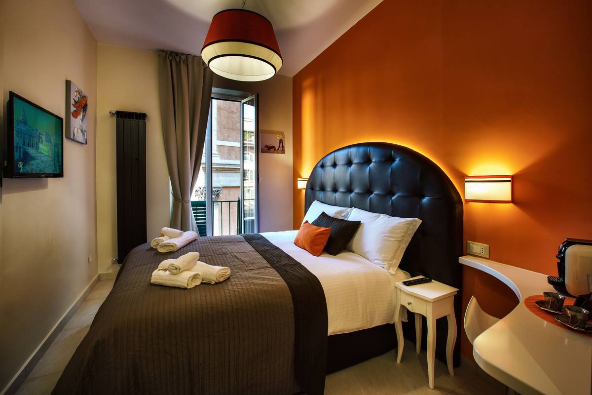 A hotel room with a large bed, a bedside table, a chair, a desk, and a view of the outside.