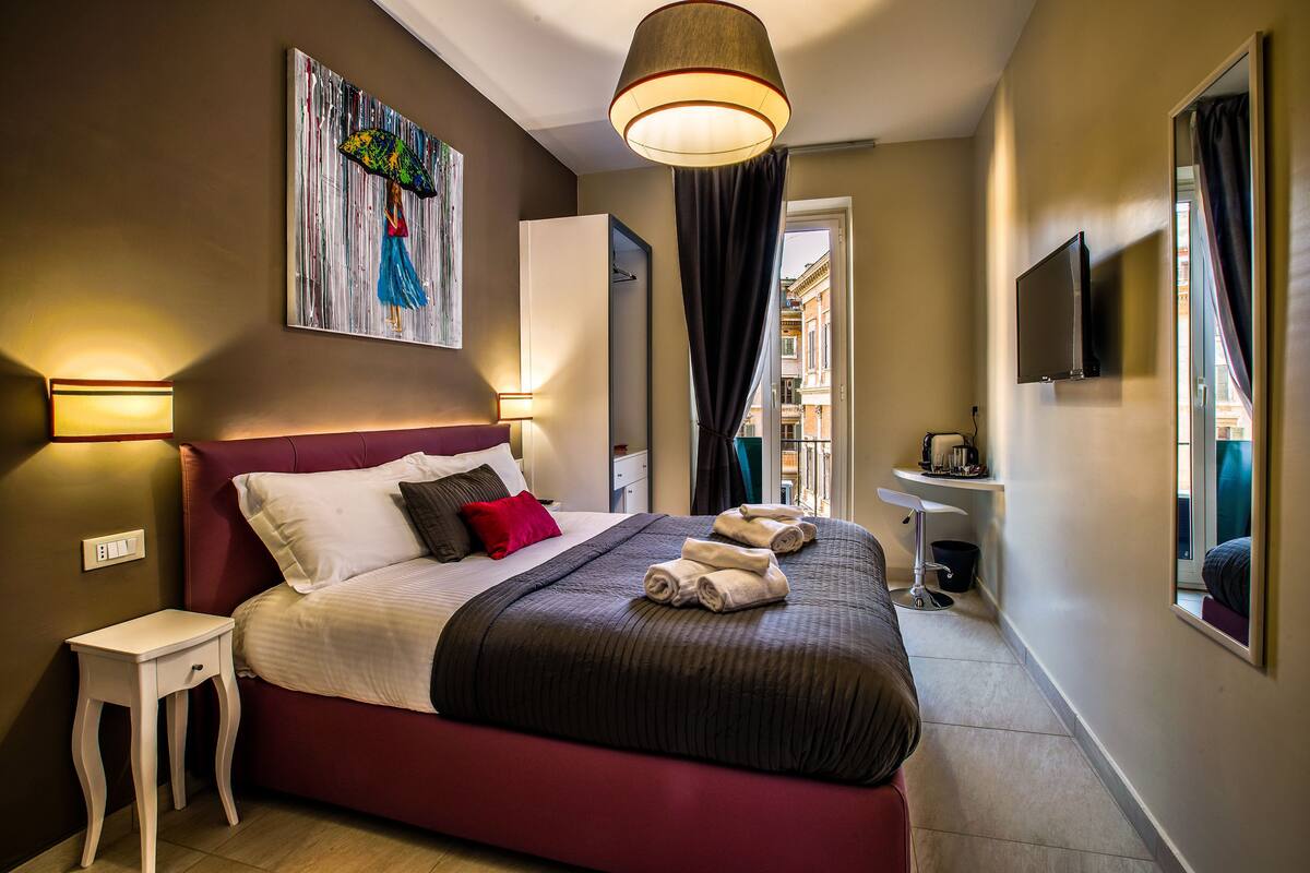 A hotel room with a large bed, a bedside table, a painting on the wall, a television, and a bathroom visible through an open door.