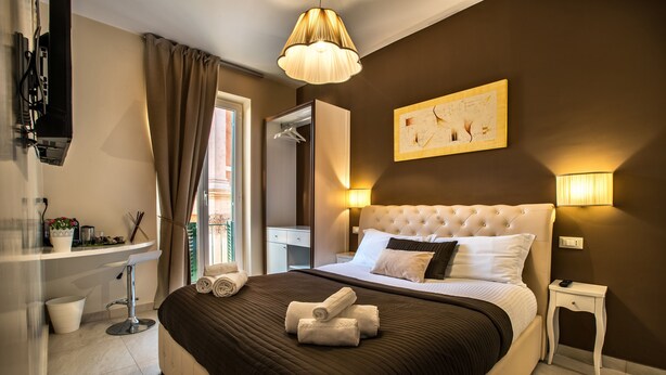A hotel room with a large bed, a bedside table, a nightstand, a mirror, a window with curtains, and a wall-mounted artwork. - Guest House Grazioli (Rome)