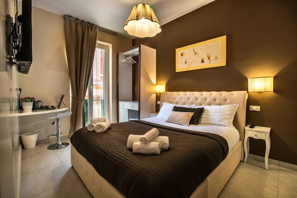 A hotel room with a large bed, a bedside table, a nightstand, a mirror, a window with curtains, and a wall-mounted artwork.