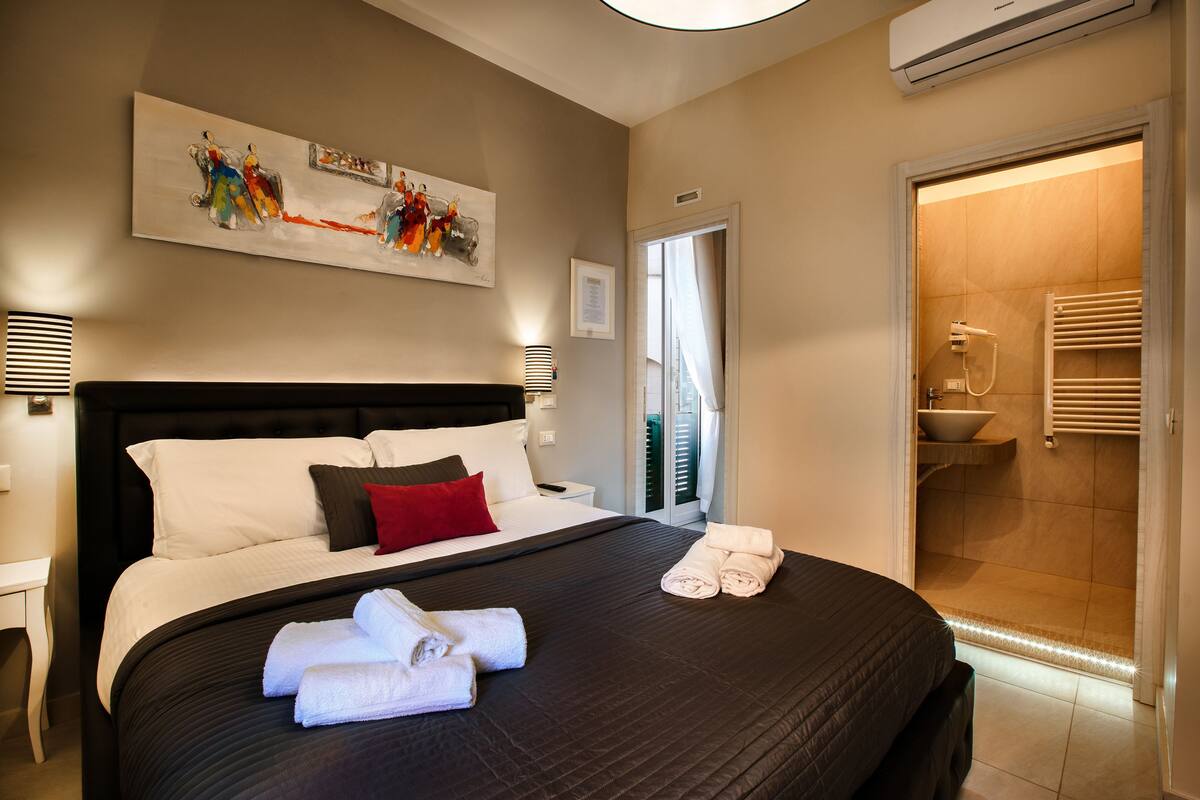 A hotel room with a large bed, a painting on the wall, a nightstand, and a bathroom with a shower and towel rack.