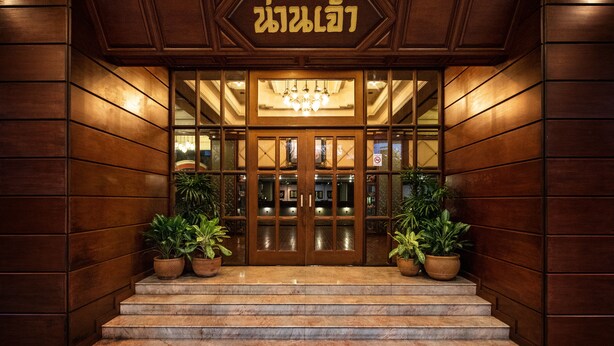 Entrada de hotel con fachada de madera, un letrero con texto, una araña y plantas en macetas.
