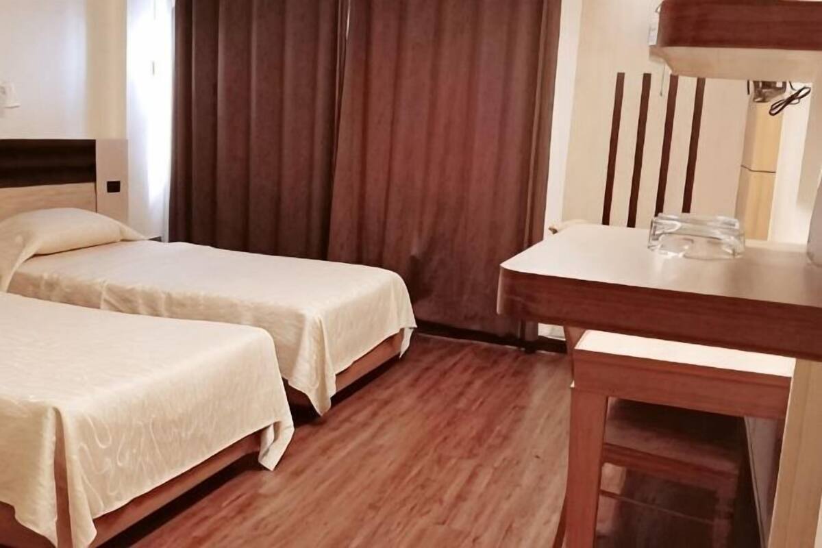 A hotel room with two single beds, a wooden desk, a chair, and a large window with curtains.