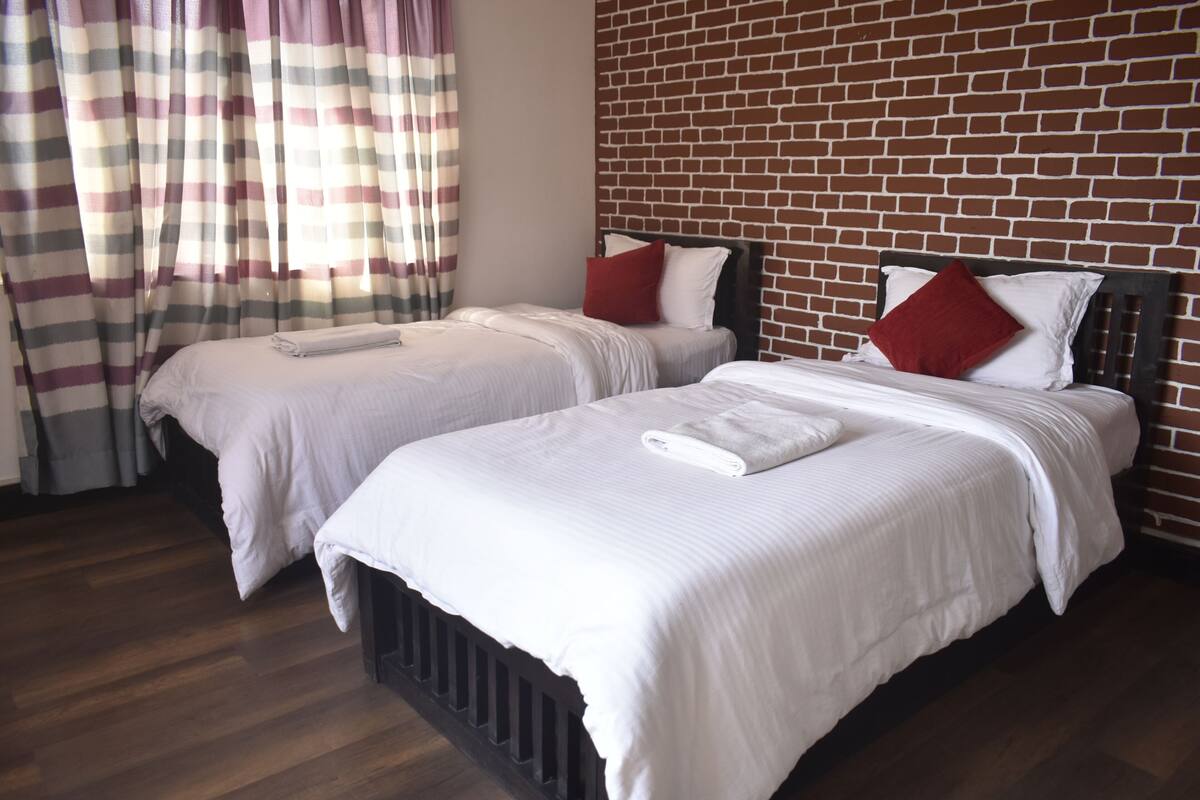 Two single beds with white linens and red pillows, a brick-patterned wall, and a window with striped curtains.