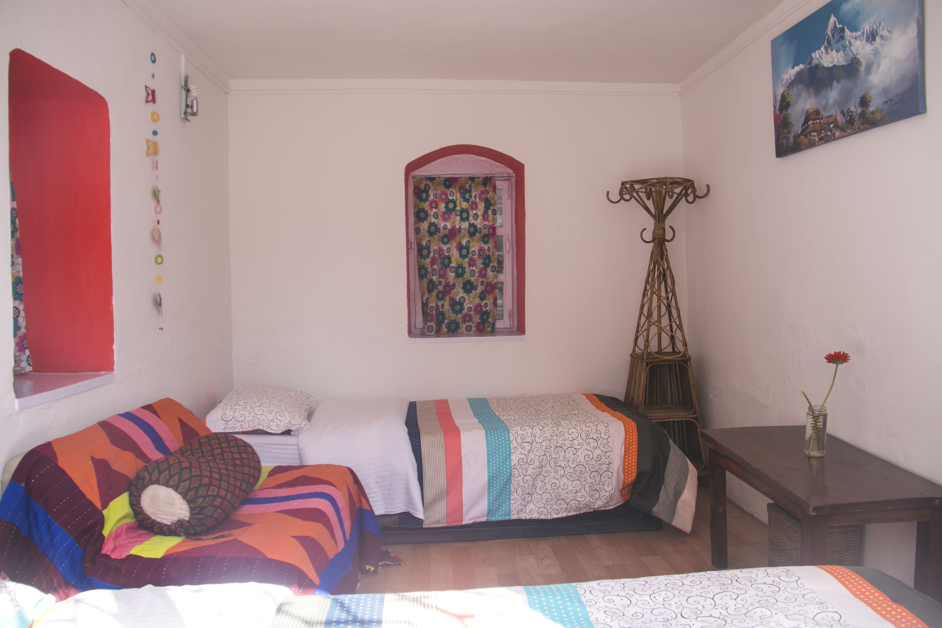 Un dormitorio con dos camas, una ventana con cortina colorida, una mesita con un florero y un cuadro en la pared.
