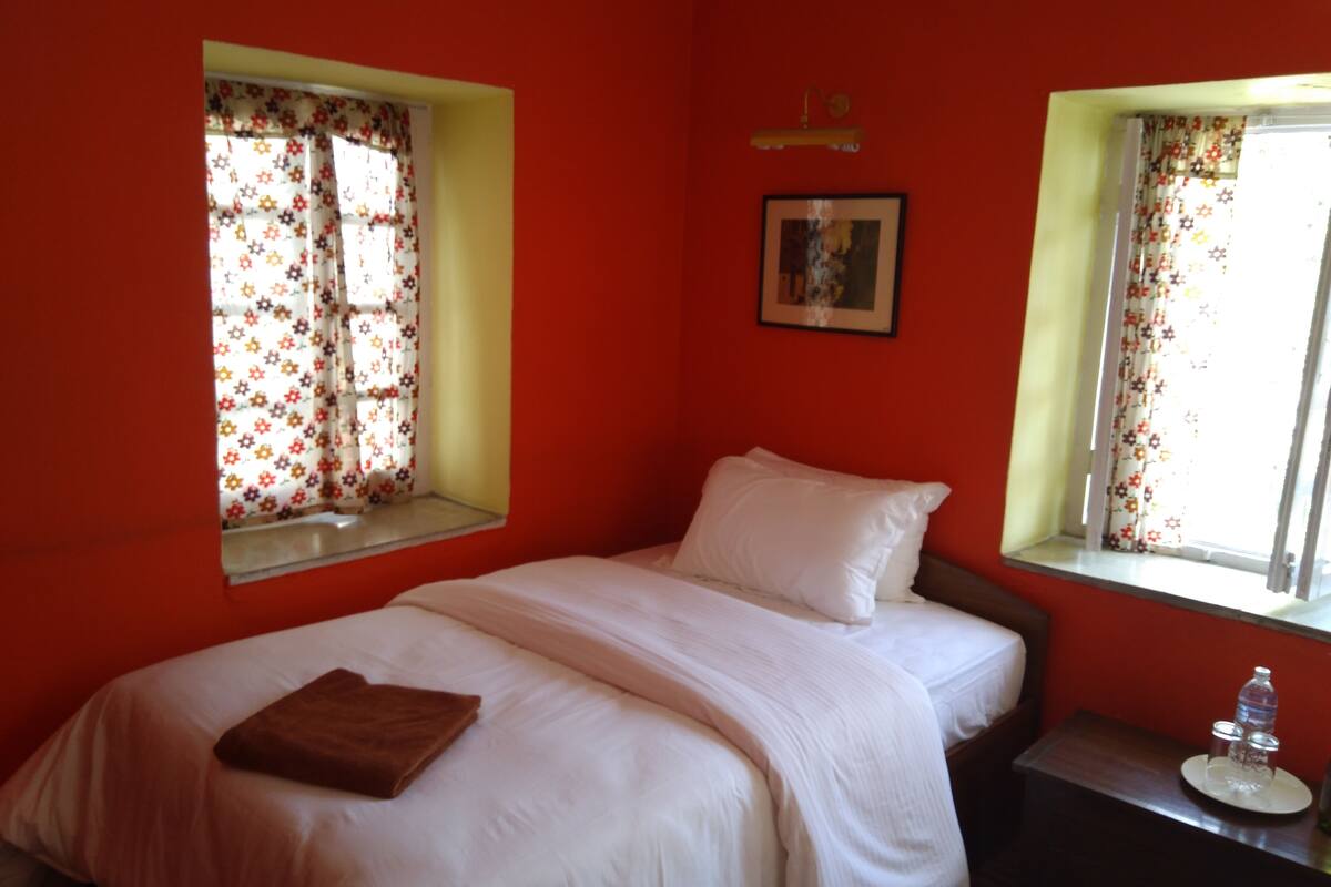 A bedroom with a bed, a nightstand, a window with floral curtains, and a wall-mounted light fixture.
