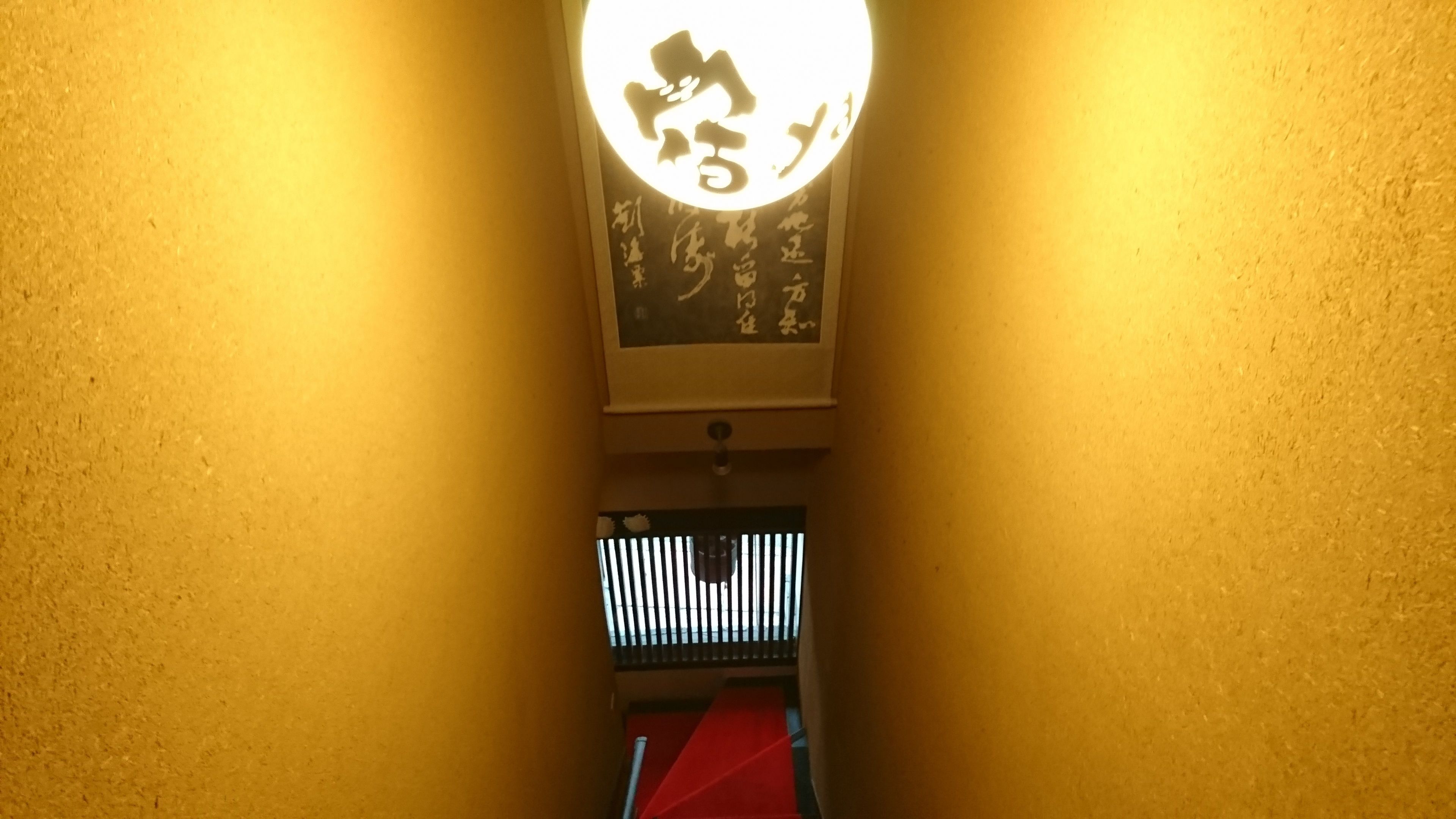 A hallway with a wall-mounted sign featuring Japanese characters and a red carpet leading to a door with vertical blinds.