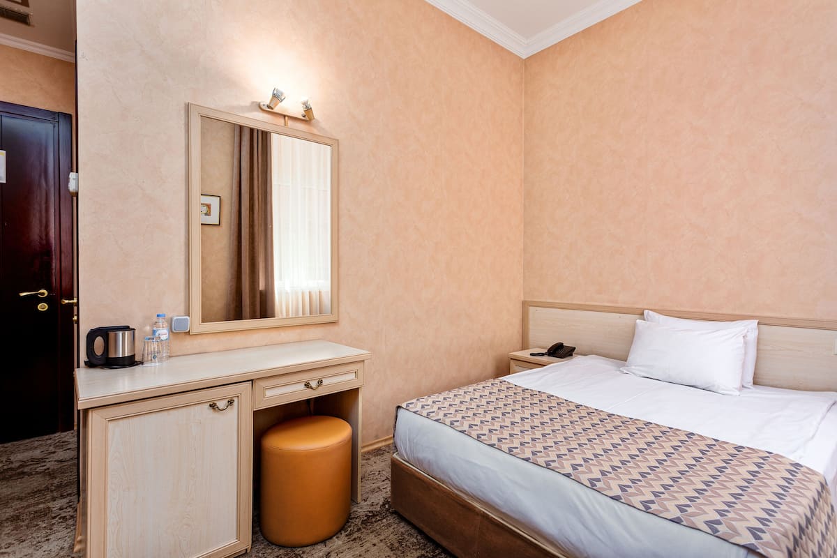 A hotel room with a bed, a bedside table, a mirror, a desk with a chair, and a small stool.