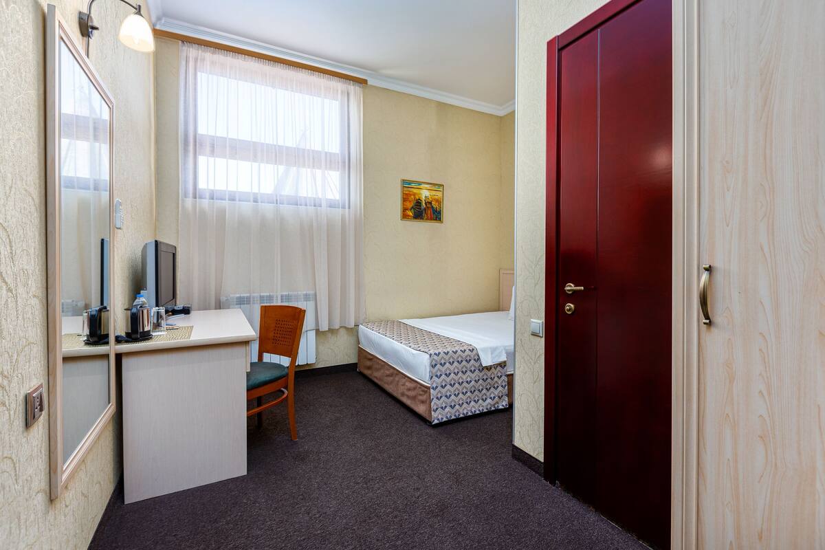 A compact hotel room with a single bed, a desk with a chair, a mirror, a small table with a kettle, and a door.