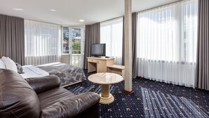 Una habitación de hotel con cama, sofá, escritorio, televisor y ventanas grandes con cortinas.