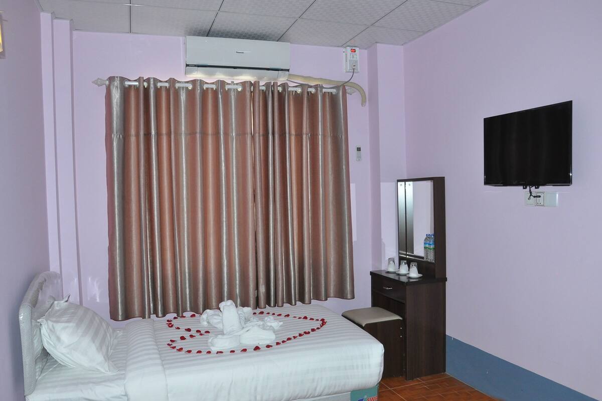 A hotel room with a bed, a desk, a mirror, a flat-screen TV, an air conditioner, and a curtain.