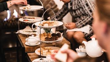 A three-tiered tea service stand with various pastries and sandwiches, a white teapot, and a cup on a wooden table.