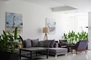 Una sala de estar moderna con un sofá gris, una mesa de centro y una lámpara. Hay dos cuadros en la pared, una planta en maceta y un ventanal grande.