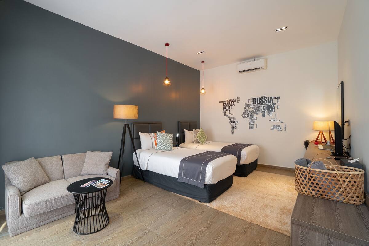 Una habitación de hotel moderna con un sofá gris, dos camas, una mesa de centro redonda y un mapa mural.