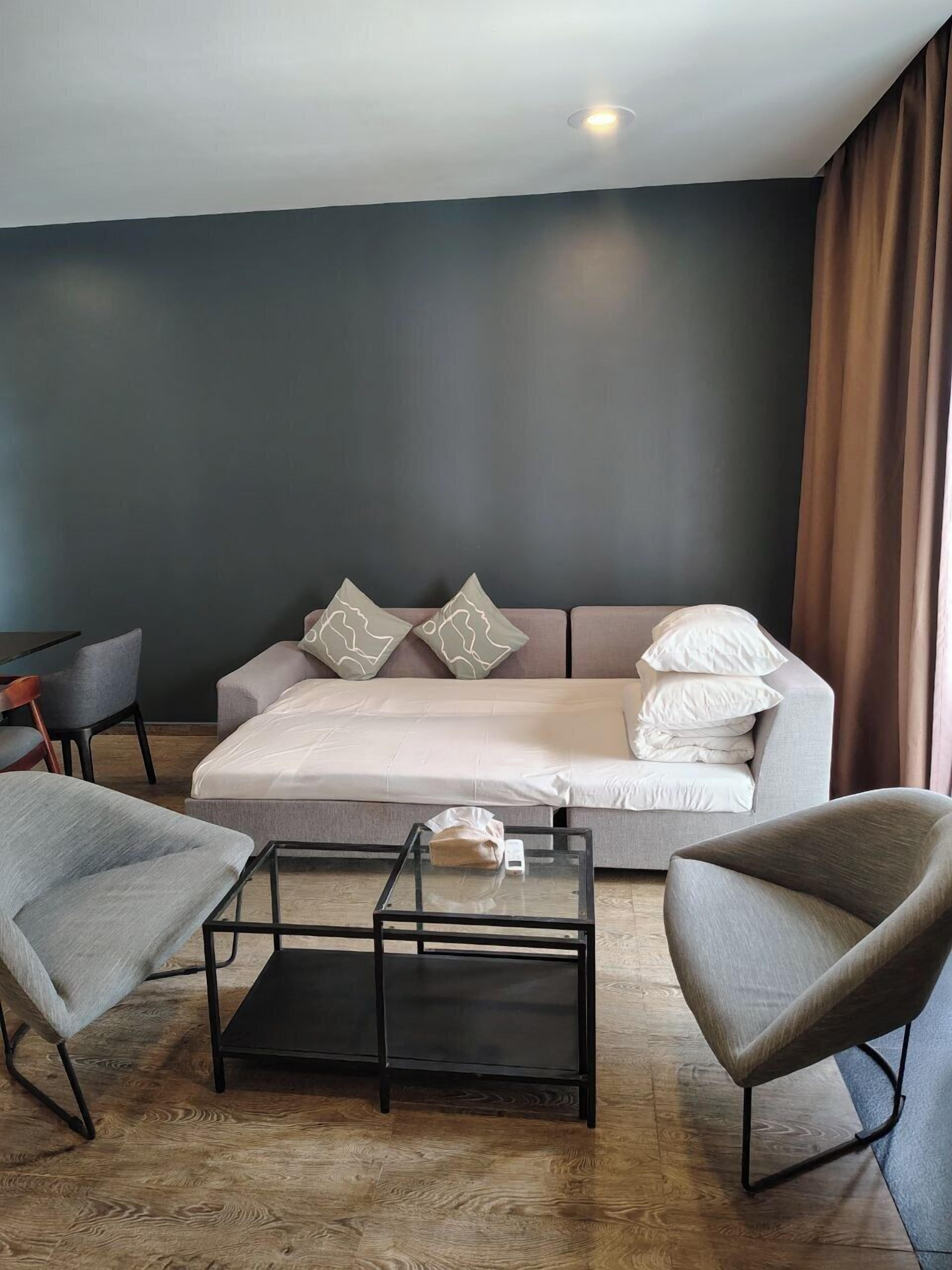 A modern hotel room with a sofa bed, a glass coffee table, and two armchairs.