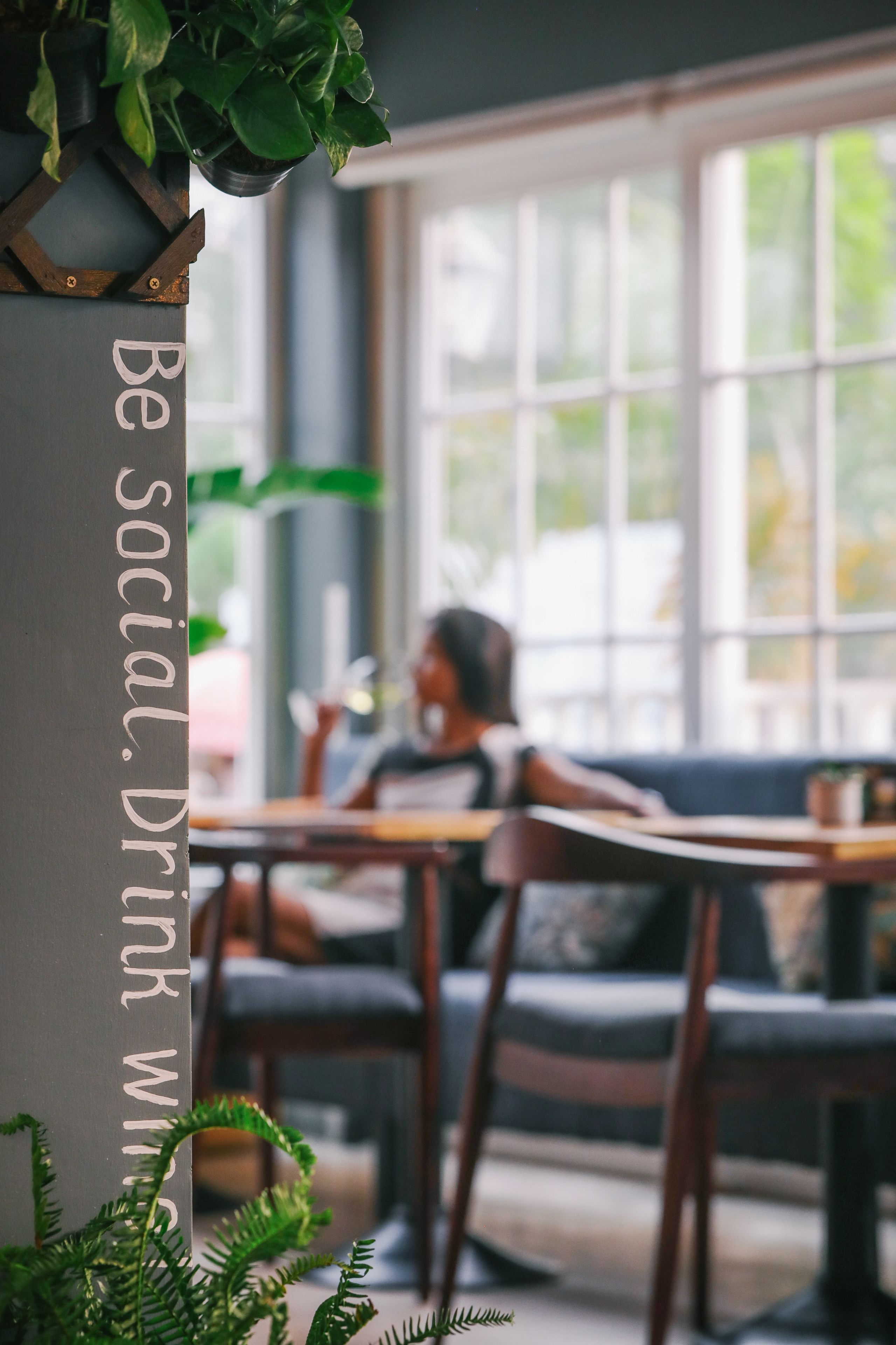 A cozy cafe interior with a large window, wooden furniture, and a sign that reads "Be social. Drink wine."