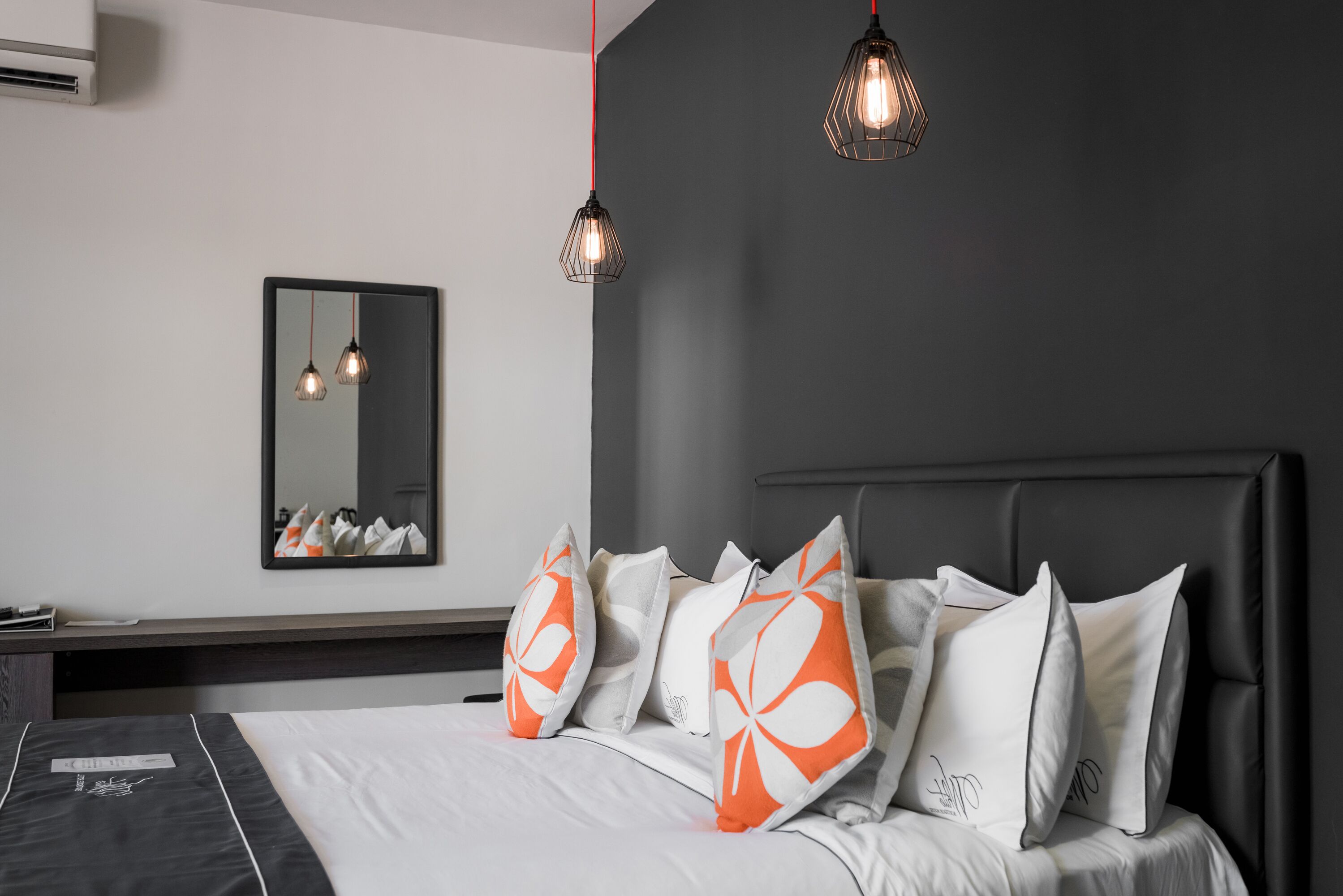 Een moderne slaapkamer met een donkergrijze muur, een groot bed met wit beddengoed en kussens met een oranje en wit patroon, en twee hanglampen.