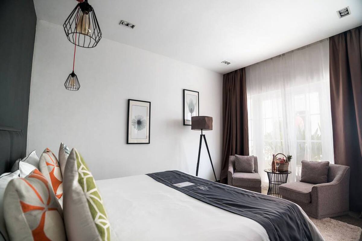 Una habitación de hotel moderna con una cama grande, dos sillones, una lámpara de pie y ventanas amplias con cortinas.