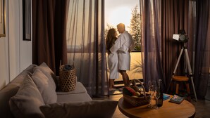 Una pareja en una habitación con un ventanal grande, un sofá, una mesa con una cesta, copas de vino y una cámara sobre un trípode.