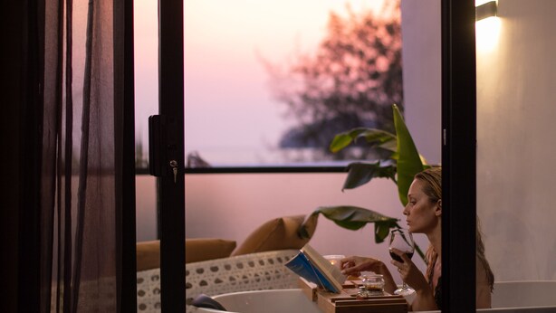 Una persona leyendo un libro en una habitación con una bañera, una planta y una ventana con vistas a árboles.