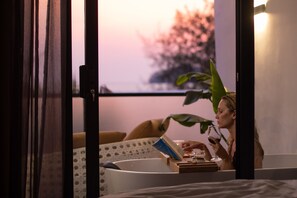 Una persona leyendo un libro en una habitación con una bañera, una planta y una ventana con vistas a árboles.