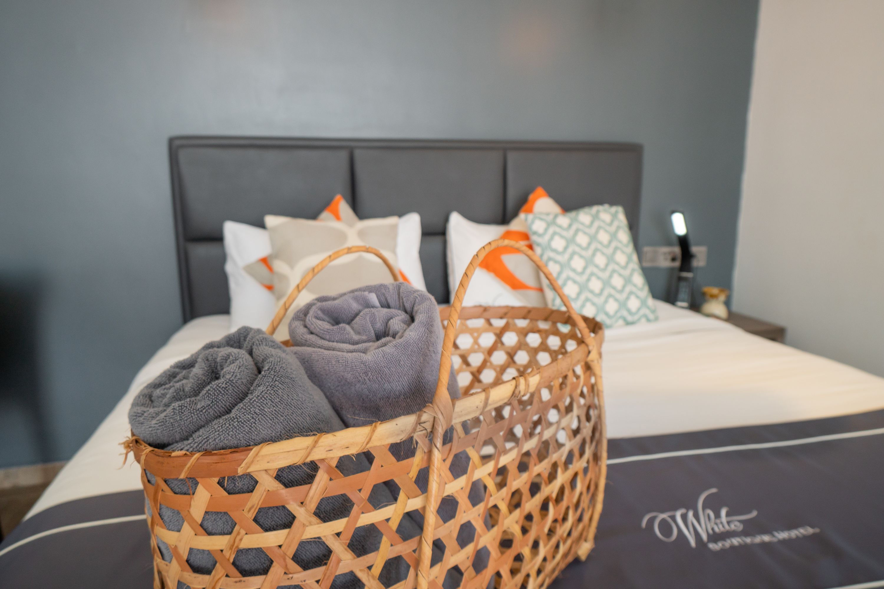 Een net opgemaakt bed met een grijs-wit gestreept laken, een grijze handdoek en een rieten mand met een grijze handdoek.