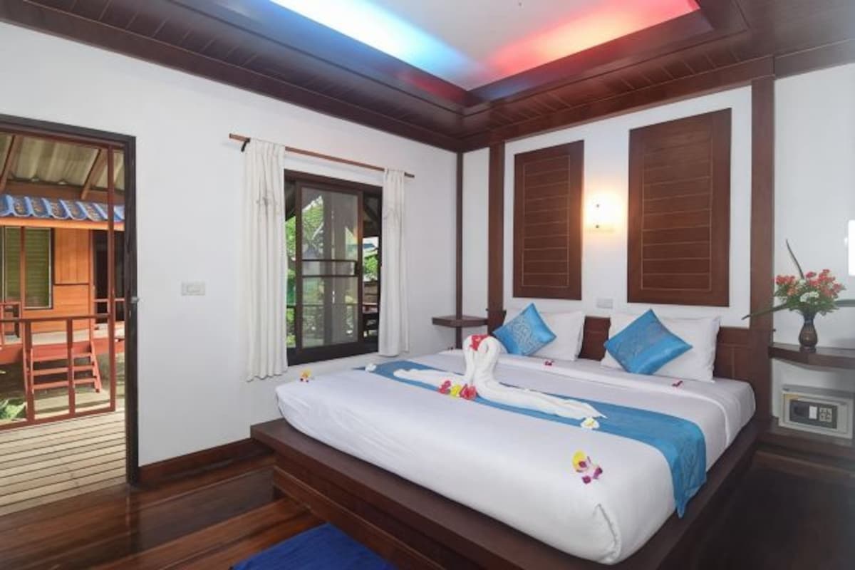 A bedroom with a bed, wooden headboard, a window with curtains, and a small table with a vase.