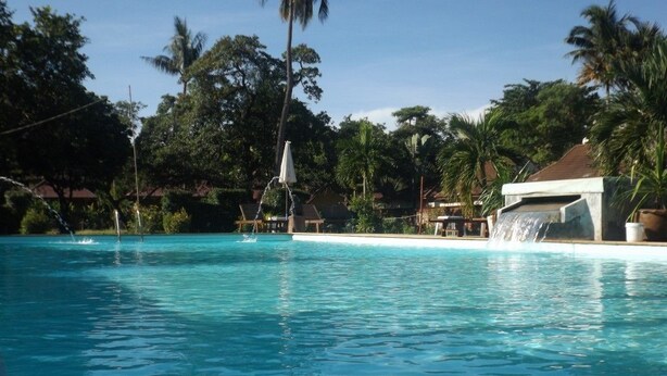 Una piscina azul cristalina con cascada, rodeada de palmeras y un edificio de techo rojo.