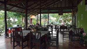 Un restaurante al aire libre con mesas y sillas de madera, cubierto por un techo de metal, rodeado de follaje.